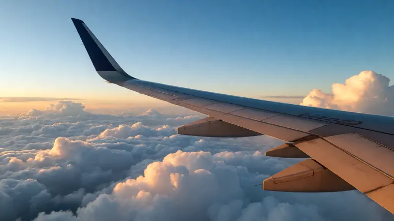 Airplane wing flying above a layer of dense white clouds under a clear blue sky during sunset.
